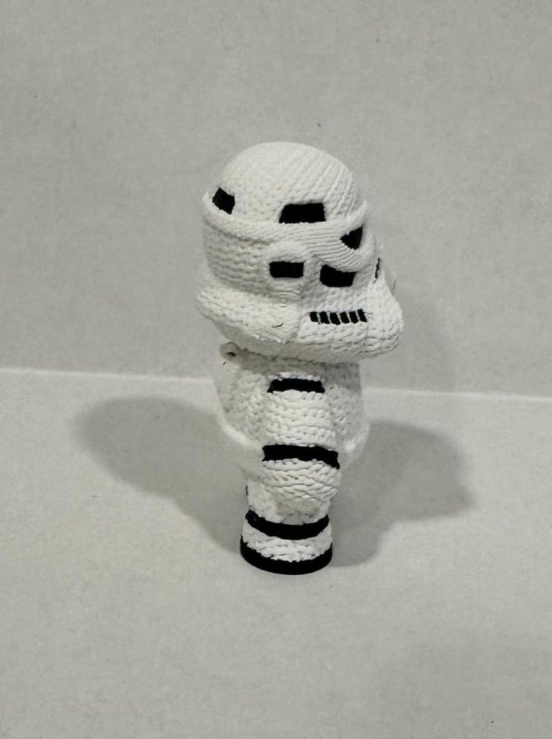 May include: White crocheted figurine of a Star Wars Stormtrooper with black details. The figurine is standing on a white surface.