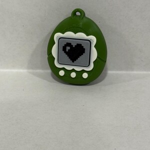 May include: A green, egg-shaped keyring featuring a pixelated heart on a grey screen. The keyring has a white scalloped border around the screen and three white dots below. A small loop is at the top.