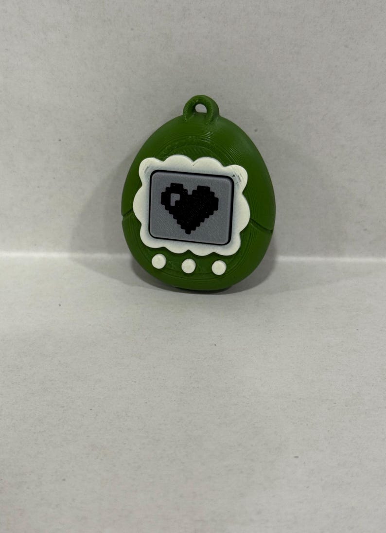 May include: A green, egg-shaped keyring featuring a pixelated heart on a grey screen. The keyring has a white scalloped border around the screen and three white dots below. A small loop is at the top.