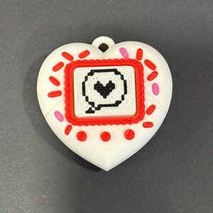 May include: A white heart-shaped pendant with a red and pink border. The center features a red square with a pixelated speech bubble containing a black heart. The pendant has a loop for hanging.