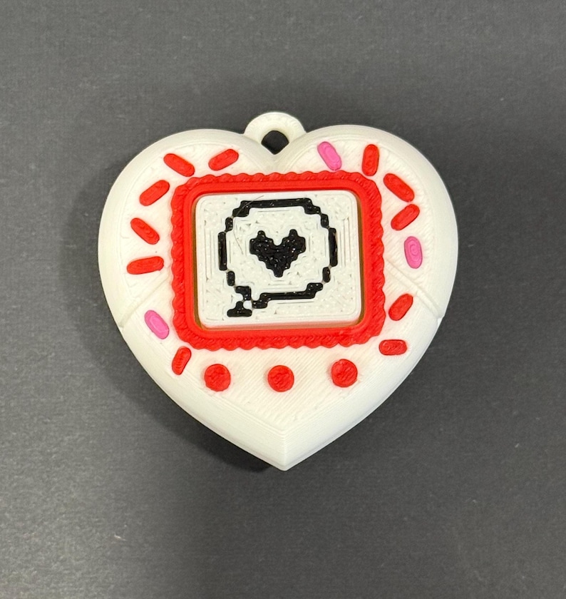 May include: A white heart-shaped pendant with a red and pink border. The center features a red square with a pixelated speech bubble containing a black heart. The pendant has a loop for hanging.