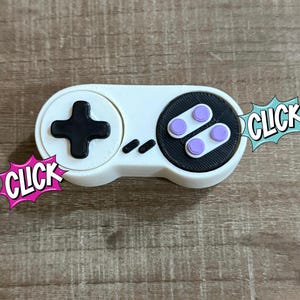 以下が含まれることがあります： 黒い十字キーと紫色のボタンが付いた白と黒のコントローラー。 コントローラーの両側に吹き出しで「CLICK」という文字が入っています。 レトロゲームアクセサリーです。