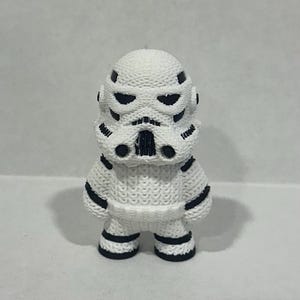 May include: A white and black stormtrooper figurine with a knitted texture. The figurine has a black face mask and black details on the body.