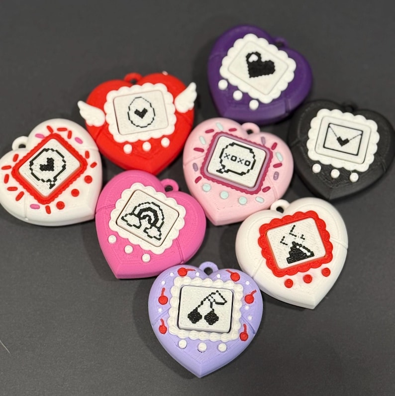 May include: A collection of heart-shaped charms in various colors, each featuring a small screen with pixelated designs. The charms include red, pink, white, purple, and black, with details like wings, rainbows, and cherries.