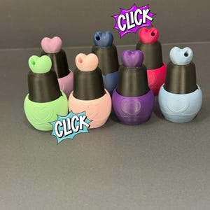 May include: A collection of colorful nail polish bottles with heart-shaped accents. Each bottle has a black cap and a different pastel color, including pink, green, purple, and blue. The word "CLICK" is displayed in a comic book style bubble.