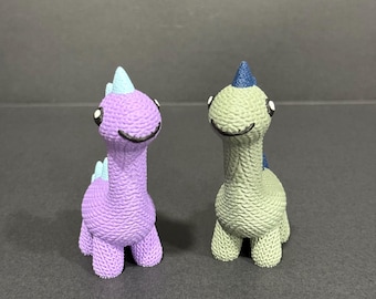 May include: Two small, textured dinosaur figurines. One is purple with light blue accents, the other is green with dark blue accents. Both have smiling faces and stand upright on four legs. The background is a solid gray.