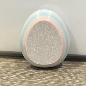May include: An egg-shaped foot file with pastel pink and light blue stripes. The centre of the file has a textured, light grey surface for exfoliation. The product is designed for foot care.