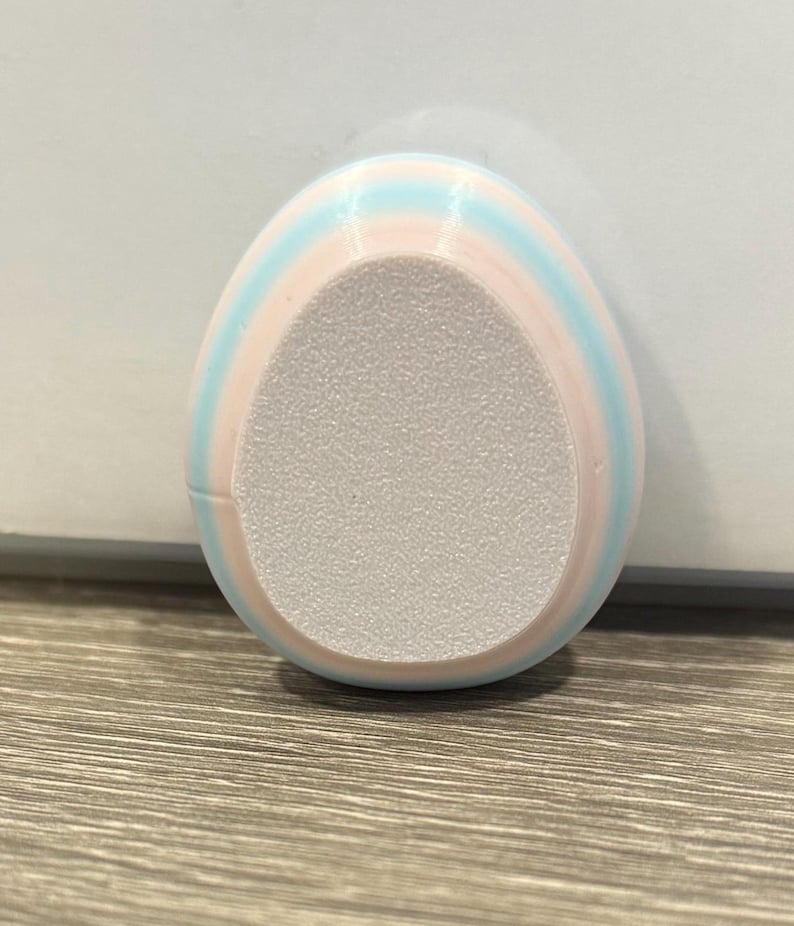 May include: An egg-shaped foot file with pastel pink and light blue stripes. The centre of the file has a textured, light grey surface for exfoliation. The product is designed for foot care.