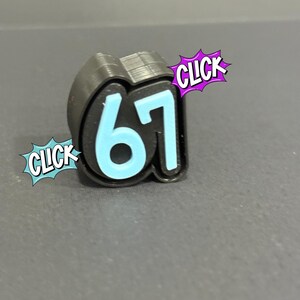 May include: A black and light blue number 67 charm with the word "CLICK" in a comic book style speech bubble. The number is light blue with a black outline. The charm is on a grey surface.