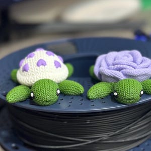 Op de afbeelding: Twee gebreide schildpadfiguren met paarse bloemen op hun schilden. De schildpadden zitten op een blauwe spoel met filament.