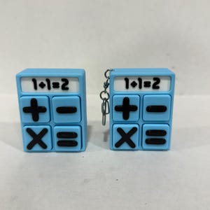 May include: Two light blue calculator-shaped keychains. Each keychain has the equation "1+1=2" printed on the top. The keychains also feature black plus, minus, multiplication, and equals signs. One keychain has a silver chain.