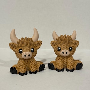 May include: Two brown knitted Highland cow figurines with black hooves and large eyes. The cows are sitting with their heads facing forward.
