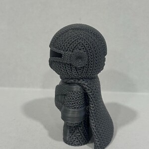May include: A gray 3D printed figurine of a character wearing a chainmail suit and a cape. The figurine is standing on a white surface.