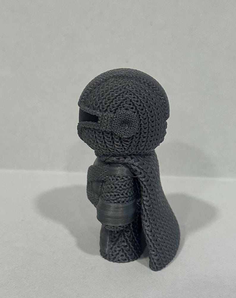 May include: A gray 3D printed figurine of a character wearing a chainmail suit and a cape. The figurine is standing on a white surface.