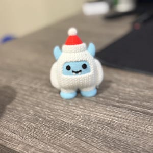 May include: A small, white crocheted yeti figure with a red and white Santa hat, blue feet, and small blue ears. The yeti has a smiling face with black eyes and a black mouth. The figure is sitting on a wooden surface.