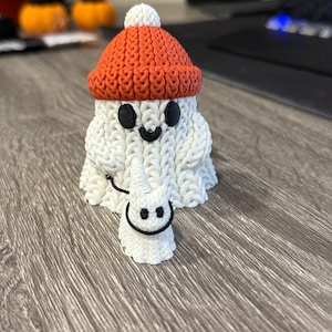以下が含まれることがあります： 笑顔の白い手作りのゴーストフィギュア。白いポンポンが付いたオレンジ色のニット帽をかぶっています。ゴーストは、笑顔の小さなゴーストフィギュアを持っています。フィギュアは木製の表面にあります。