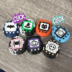 Puede incluir: Una colección de siete accesorios de juguete con temática de Halloween en varios colores, incluyendo azul, naranja y negro. Cada juguete presenta un icono espeluznante diferente, como un murciélago, un sombrero de bruja o un fantasma, dentro de un marco blanco. La palabra "CLICK" se muestra.