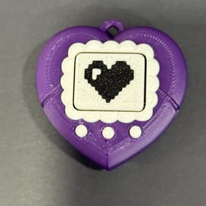 May include: A purple heart-shaped handheld game device with a white screen and a black pixelated heart design. The device has three white buttons below the screen and a loop at the top for attaching a lanyard.