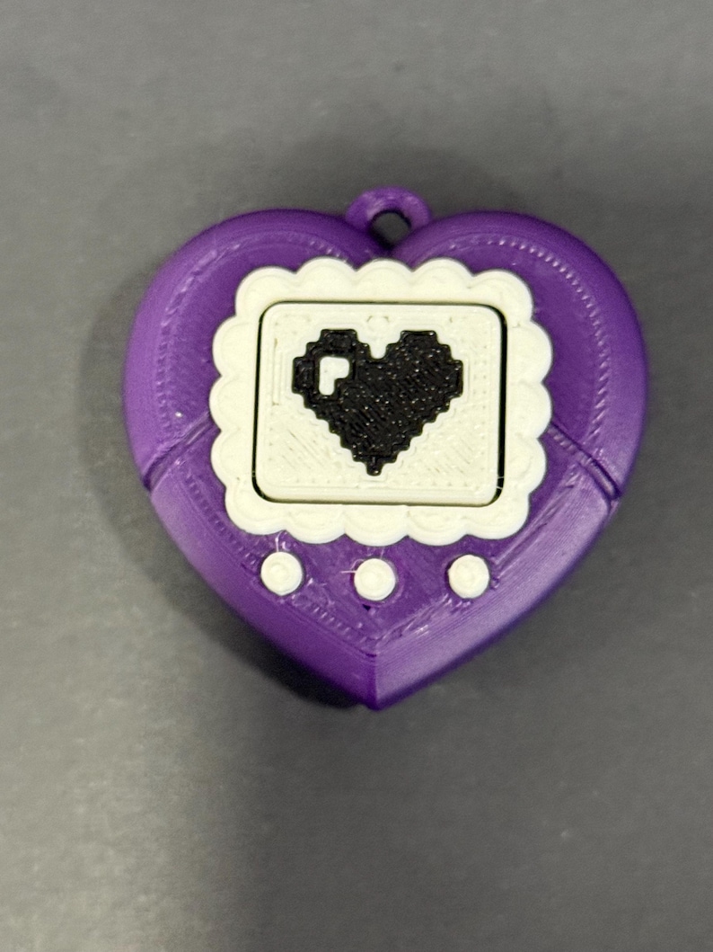 May include: A purple heart-shaped handheld game device with a white screen and a black pixelated heart design. The device has three white buttons below the screen and a loop at the top for attaching a lanyard.