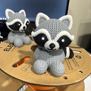 May include: Two gray knitted raccoon figurines with black eyes and white accents. The figurines are sitting on a spool of orange filament.