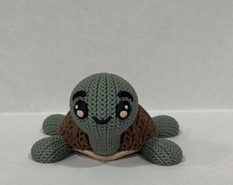Peut inclure: Une petite tortue en tricot vert avec des accents bruns et un visage souriant. La tortue a de grands yeux noirs avec des pupilles blanches.
