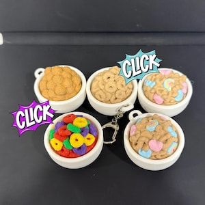 May include: A collection of miniature cereal bowls, each filled with different types of cereal. The bowls are white with a handle. The cereal includes brown, colorful, and frosted varieties. The word "CLICK" is in a speech bubble.