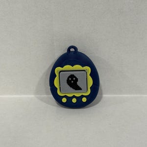 May include: A blue digital pet toy with a yellow border and a pixelated black ghost on the screen. It has three yellow buttons and a loop. The toy is a retro design.