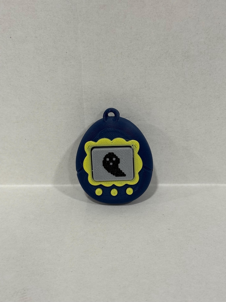 May include: A blue digital pet toy with a yellow border and a pixelated black ghost on the screen. It has three yellow buttons and a loop. The toy is a retro design.