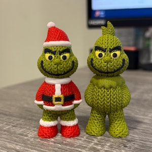 May include: Two green figurines of a Christmas character. One is dressed in a red Santa suit with a black belt and gold buckle, and a red hat. The other is in a green sweater. Both have yellow eyes and a stitched appearance.
