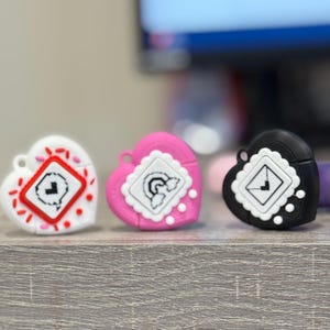 May include: Three heart-shaped cases in white, pink, and black. Each case features a square design with a unique graphic. The white case has red accents, the pink case has a white scalloped border, and the black case has white dots.