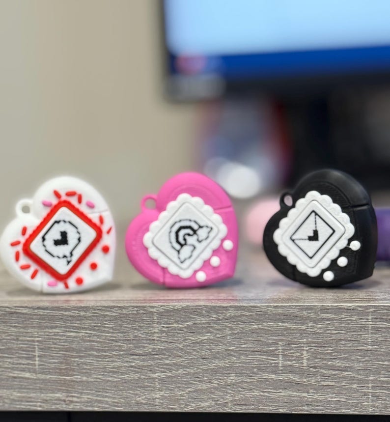 May include: Three heart-shaped cases in white, pink, and black. Each case features a square design with a unique graphic. The white case has red accents, the pink case has a white scalloped border, and the black case has white dots.