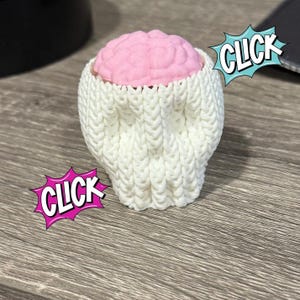May include: A pink brain-shaped object sits inside a white knitted cozy. The word "CLICK" is displayed in a speech bubble on the image. The cozy is made of thick, textured yarn.