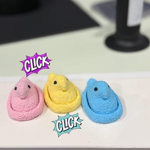 May include: Three colorful marshmallow chick candies in pink, yellow, and blue. Each chick has black dot eyes. The word "CLICK" is in a comic book style speech bubble next to the pink and blue chicks.