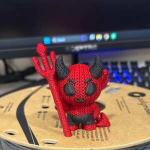 May include: A red and black 3D printed figurine of a devil holding a trident. The devil has a knitted texture and a mischievous expression.