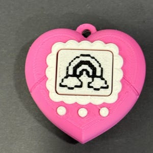 May include: A pink heart-shaped pendant with a white scalloped frame around a small screen displaying a black and white rainbow design. The pendant has three white circular buttons below the screen and a small loop at the top.