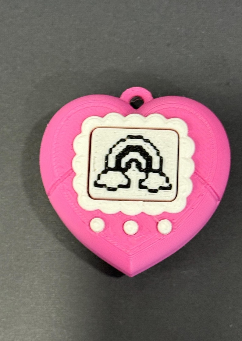 May include: A pink heart-shaped pendant with a white scalloped frame around a small screen displaying a black and white rainbow design. The pendant has three white circular buttons below the screen and a small loop at the top.
