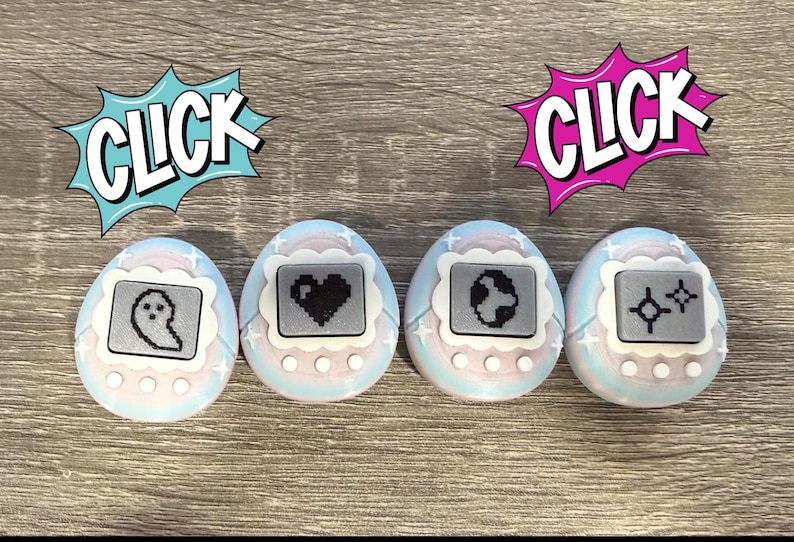 May include: Four pastel-coloured digital pet toys with pixelated icons of a ghost, heart, globe, and stars. The toys have a white border and are set against a wooden background. Comic book style speech bubbles with the word "CLICK" are above two of the toys.