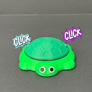 May include: A bright green turtle-shaped container with a textured shell and a smiling face. The word "CLICK" is displayed in a comic book style speech bubble on either side of the turtle.