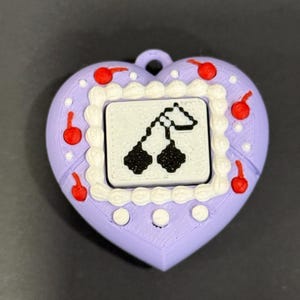 May include: A heart-shaped pendant in lavender with a white border and red cherries. The center features a small screen with a black and white pixelated cherry design. The pendant has a loop for attaching to a chain or keychain.