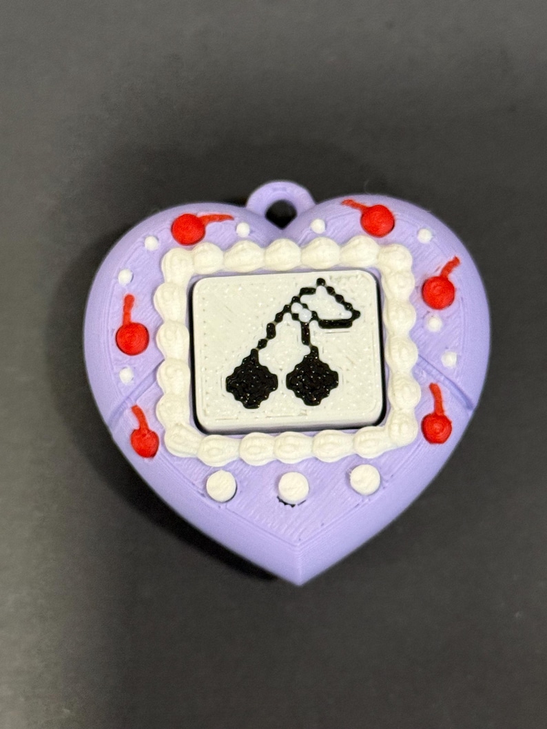 May include: A heart-shaped pendant in lavender with a white border and red cherries. The center features a small screen with a black and white pixelated cherry design. The pendant has a loop for attaching to a chain or keychain.