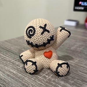 以下が含まれることがあります： 黒い縫い目のディテールと赤いハートが付いたベージュ色のブードゥー人形。人形には縫い付けられた口、目のためのX字型のステッチ、片方の目のスパイラルステッチがあります。人形は木製の表面に座っています。