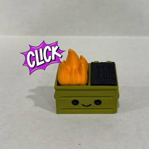 Op de afbeelding: Een groene miniatuurvuilcontainer met een lachend gezicht, een zwarte grill en oranje vlammen. Het woord "CLICK" staat in een paarse tekstballon. De container is een grillig, decoratief item.