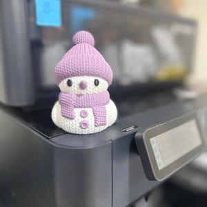 May include: A small purple and white snowman figurine with a knitted hat and scarf. The snowman is sitting on top of a black printer.