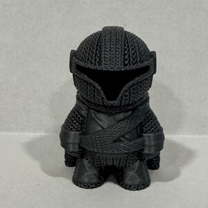 May include: A black 3D printed figurine of a knight wearing chainmail armor and a helmet. The figurine is standing with its arms crossed.
