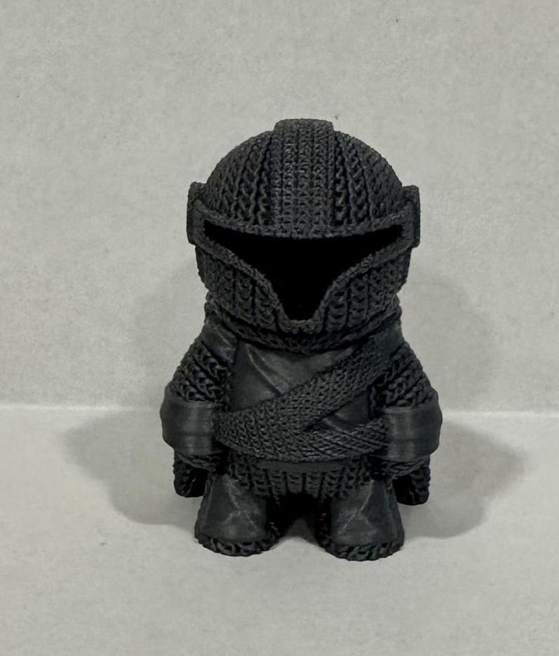 May include: A black 3D printed figurine of a knight wearing chainmail armor and a helmet. The figurine is standing with its arms crossed.