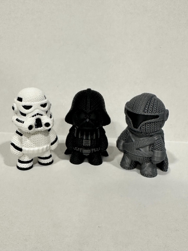 May include: Three knitted Star Wars characters: a white Stormtrooper, a black Darth Vader, and a gray Mandalorian. The characters are all standing on a white surface.