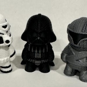 May include: Three small, intricately detailed, 3D printed figurines of Star Wars characters. The characters are Darth Vader, a Stormtrooper, and a Mandalorian. The figurines are all black and white.
