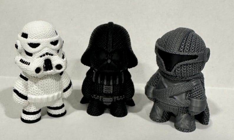 May include: Three small, intricately detailed, 3D printed figurines of Star Wars characters. The characters are Darth Vader, a Stormtrooper, and a Mandalorian. The figurines are all black and white.