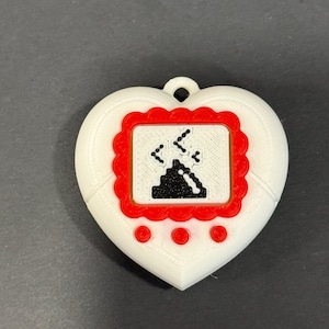 May include: A white heart-shaped pendant with a red-framed screen displaying a pixelated graphic. The screen shows a black object with falling lines. Three red circles are below the screen. A small loop is at the top.