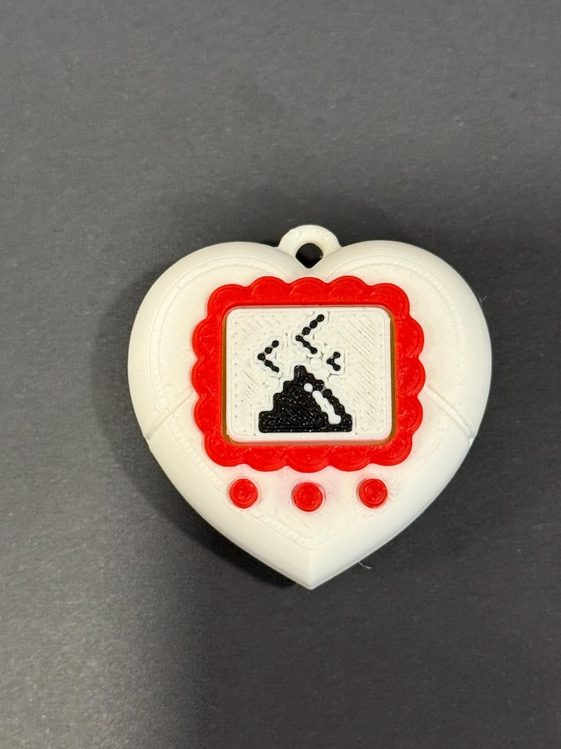 May include: A white heart-shaped pendant with a red-framed screen displaying a pixelated graphic. The screen shows a black object with falling lines. Three red circles are below the screen. A small loop is at the top.